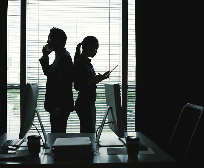 Two professional silhouettes standing back to back in an office, one on a phone and one on a tablet