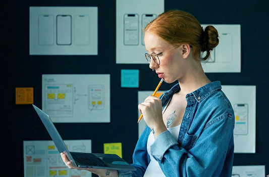 Woman holding a laptop in front of a wall with UI wireframes and sticky notes